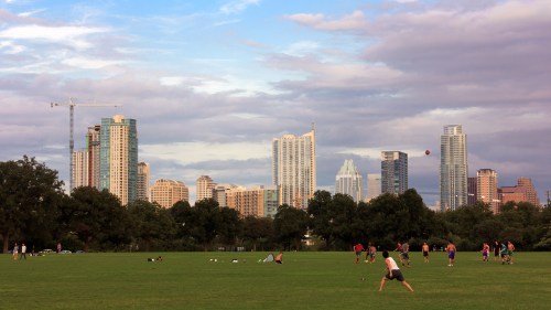 A city skyline with tall buildings in the background, people playing sports on a green field in the foreground.