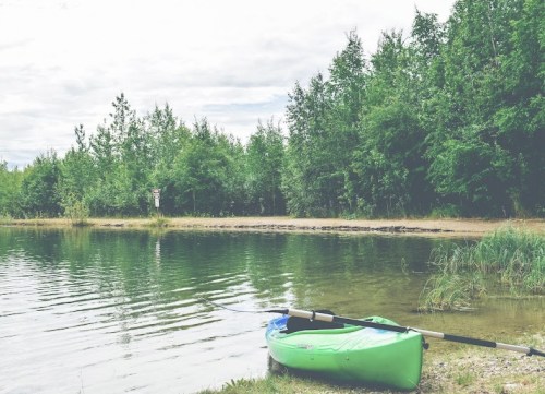 A green kayak rests on the shore of a calm lake, surrounded by lush greenery and a cloudy sky.