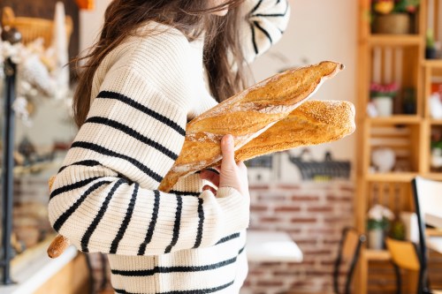 A person in a striped sweater holds two fresh baguettes in a cozy bakery setting.