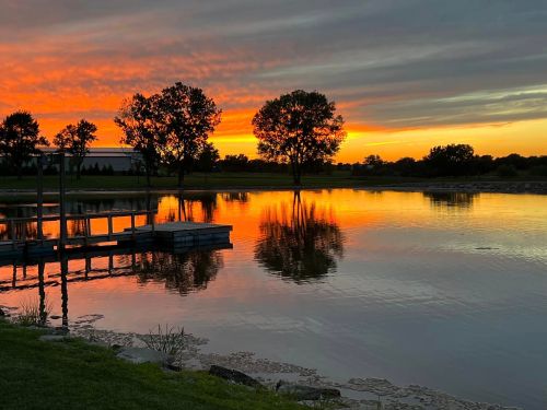 A serene lake at sunset, with vibrant orange and purple skies reflecting on the water and silhouetted trees in the background.