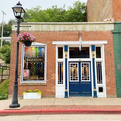 A brick building with a blue door and colorful bakery sign, featuring hanging flower baskets and a streetlamp.