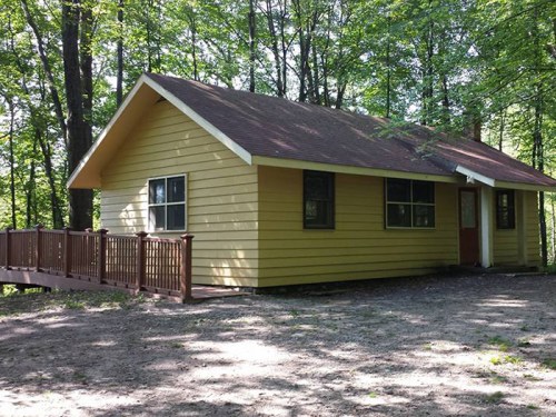 A yellow house with a brown roof surrounded by trees, featuring a wooden deck and gravel path.