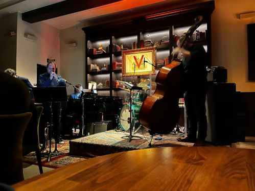 A jazz band performs on stage, featuring a pianist, saxophonist, and double bassist, with a cozy ambiance.