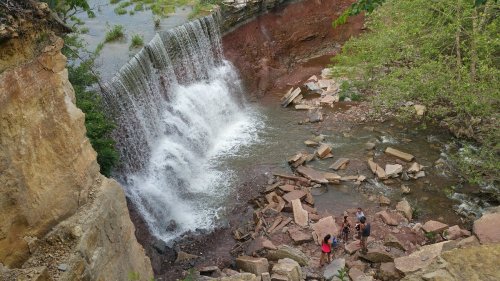 A waterfall cascades over rocky cliffs, with four people exploring the area below amidst lush greenery.
