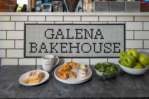 A cozy bakery display featuring pastries, a salad, a coffee mug, and green apples against a tiled backdrop.