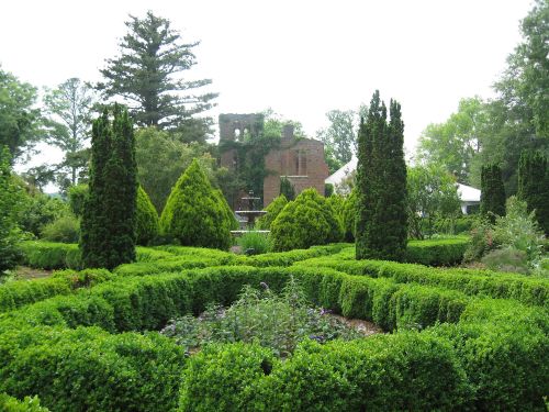 Lush green garden with neatly trimmed hedges, conical trees, and a historic brick building in the background.