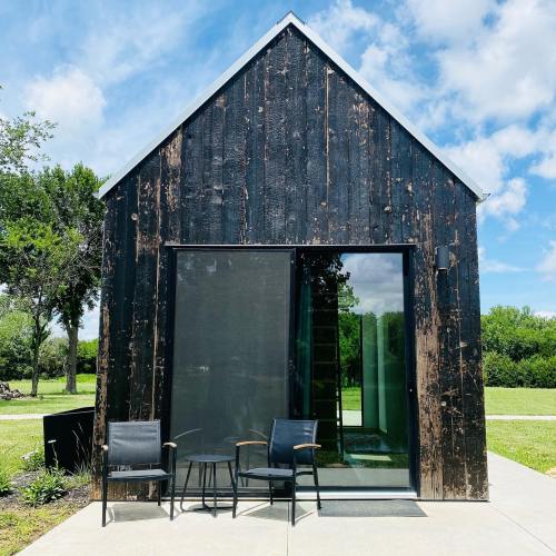 A modern black wooden cabin with a glass door, flanked by two chairs, set against a blue sky and green landscape.