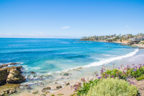 A scenic view of a beach with clear blue water, gentle waves, and coastal cliffs under a bright blue sky.