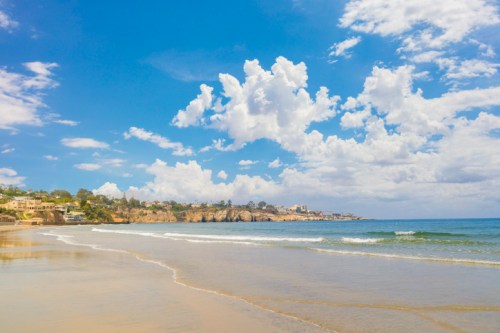 A serene beach scene with gentle waves, sandy shore, and a bright blue sky dotted with fluffy white clouds.