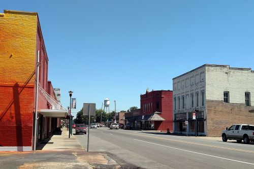 A sunny street view of a small town with colorful buildings, a water tower in the distance, and clear blue skies.