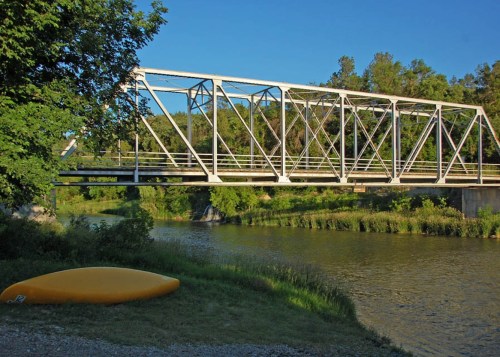 A white metal bridge spans a calm river, with a yellow kayak resting on the grassy bank under a clear blue sky.