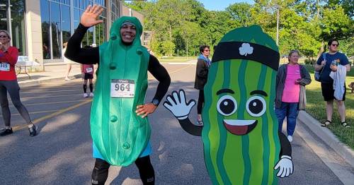 A person in a pickle costume poses with a cartoon pickle cutout at a fun run event, with participants in the background.