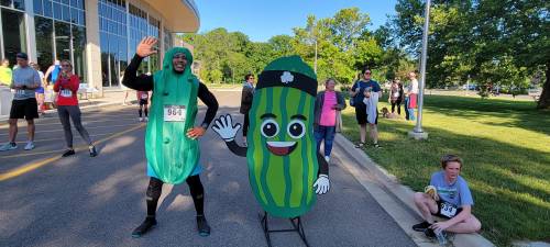 A person in a pickle costume waves next to a cartoon pickle cutout at a fun run event, with participants in the background.