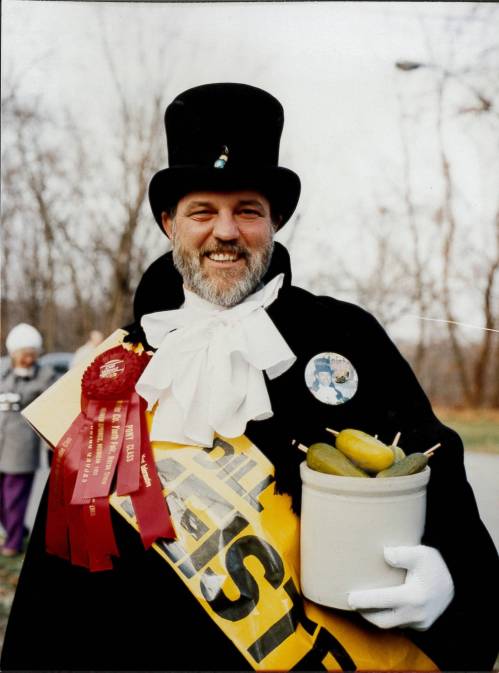 A smiling man in a top hat and cape holds a jar of pickles, wearing ribbons and a badge at an outdoor event.