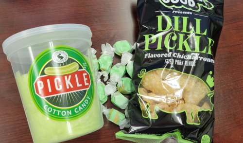 A container of pickle-flavored cotton candy next to a bag of dill pickle-flavored pork rinds and green candies.