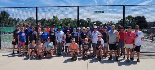 A large group of people poses together on a tennis court, smiling under a clear blue sky.