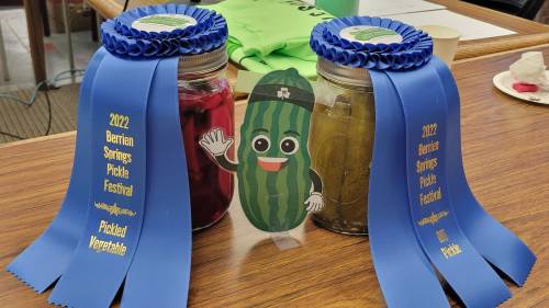 Two jars of pickled vegetables with blue ribbons from the 2022 Berrien Springs Pickle Festival and a cartoon pickle mascot.