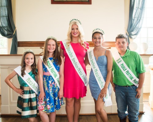 Five children pose together, wearing crowns and sashes for the Pickle Festival, in a bright, cheerful setting.