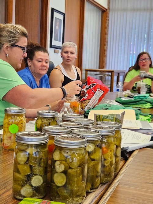 Four women sit around a table with jars of pickles in the foreground, engaged in conversation and enjoying snacks.
