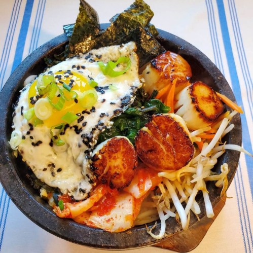 A stone bowl filled with rice, topped with a fried egg, scallions, seaweed, scallops, and assorted vegetables.