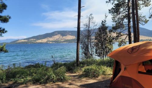 A vibrant orange tent by a serene lake, surrounded by trees and mountains under a clear blue sky.