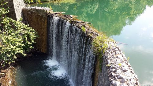 A serene waterfall cascading over a stone wall into a calm, green-tinted pool surrounded by lush vegetation.