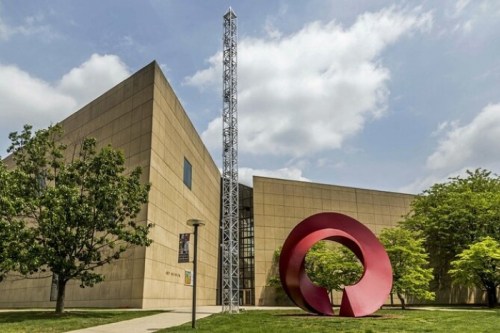 A modern building with a large red sculpture in front, surrounded by trees and a cloudy sky.