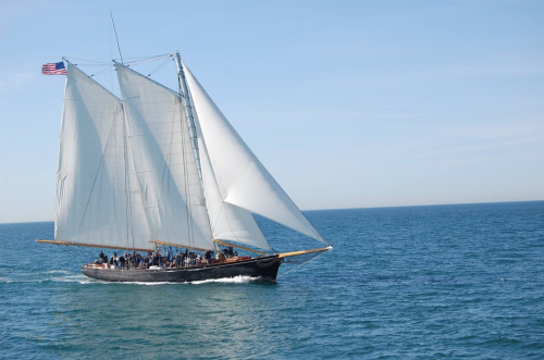 A tall ship with white sails sailing on blue waters, with people visible on deck and an American flag flying.