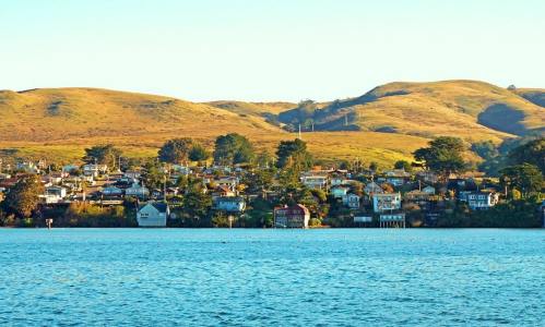 A serene waterfront view of a small town with houses nestled against rolling hills and clear blue water.