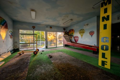 Abandoned mini golf course with colorful hot air balloon murals and a view of the sunset through large windows.