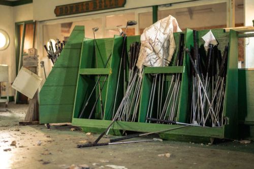 A cluttered rack of golf clubs in a green wooden storage area, with a few scattered items on the floor.