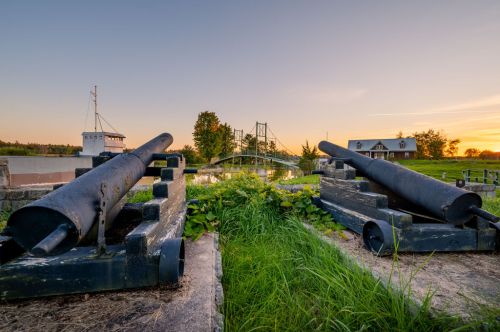Two historic cannons on display, surrounded by grass, with a bridge and buildings in the background at sunset.