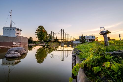 A serene landscape featuring a bridge over calm water, with a ship and trees reflecting in the sunset-lit scene.
