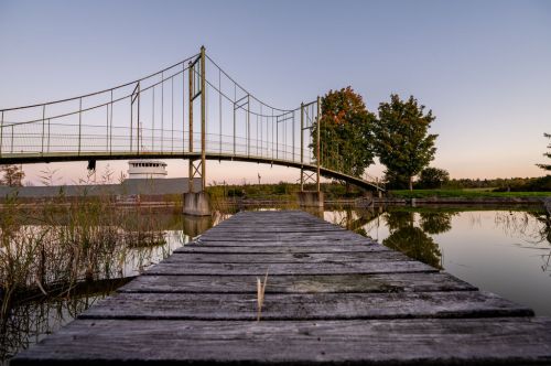 A wooden dock leads to a bridge over calm water, surrounded by trees at sunset.
