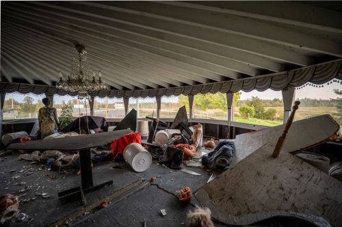 An abandoned room with scattered debris, broken furniture, and a chandelier, overlooking a scenic view outside.