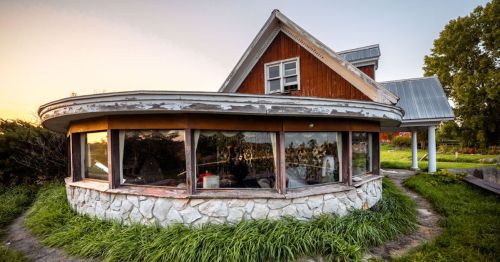 A unique, round house with a wooden upper section and stone base, surrounded by lush green grass at sunset.