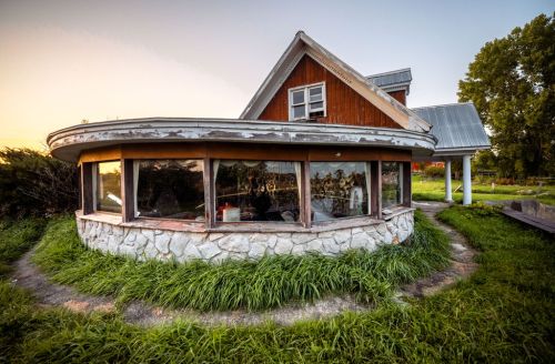 A rustic, round house with a stone base and large windows, surrounded by tall grass at sunset.