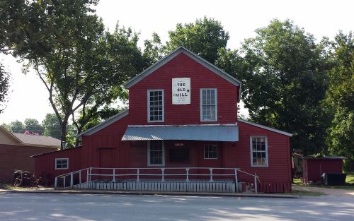 A red wooden building with a sign reading "The Old Mill," surrounded by trees and a clear blue sky.