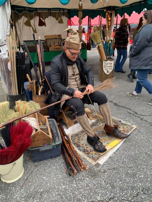 A man in period clothing sits at a market stall, crafting brooms and sticks, surrounded by various handmade items.