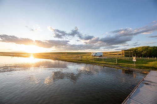 A serene landscape at sunset, featuring a calm lake, grassy shore, and a van parked nearby under a cloudy sky.