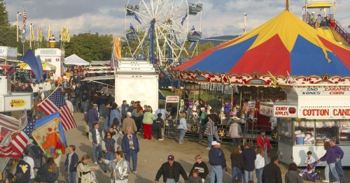 A bustling fairground scene with a Ferris wheel, colorful tents, food stalls, and crowds enjoying the festivities.