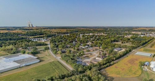 Aerial view of a small town surrounded by greenery and fields, with industrial structures in the background.