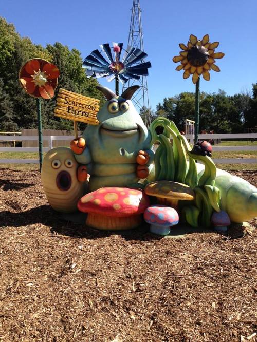 Colorful cartoon-like sculptures of a monster and mushrooms in a playful farm setting with flowers and a windmill.