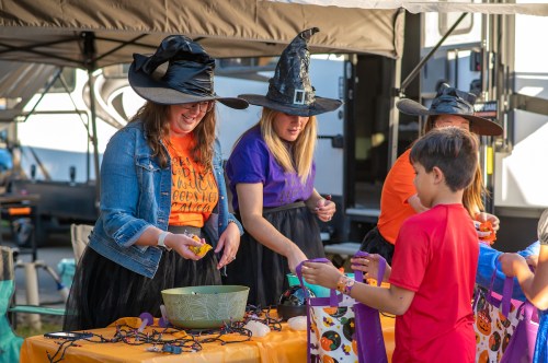Two women in witch hats and costumes assist children at a Halloween-themed table with decorations and treats.