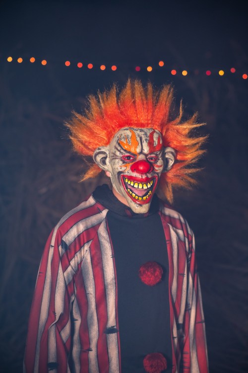 A creepy clown with bright orange hair, a painted face, and a striped costume stands in a dimly lit setting.