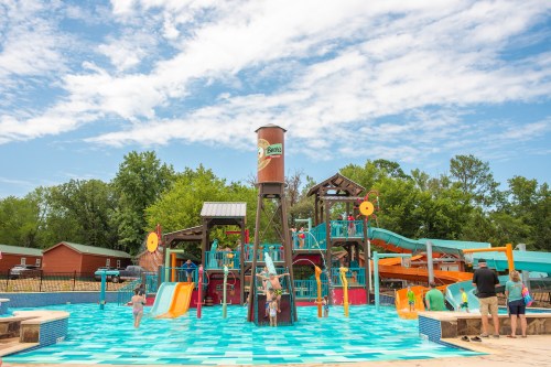 A colorful water play area with slides, a splash tower, and families enjoying the pool on a sunny day.