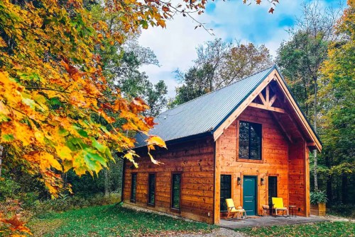 A cozy wooden cabin surrounded by colorful autumn leaves and trees under a blue sky.