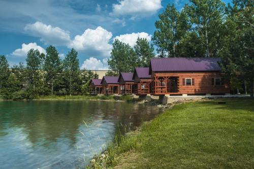 Log cabins by a serene pond, surrounded by lush greenery and a blue sky with fluffy clouds.