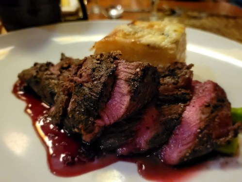A plate featuring sliced, medium-rare steak with a rich sauce and a side of baked dish.