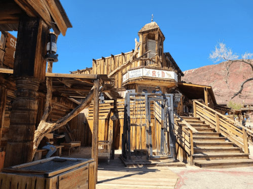 A rustic wooden building with stairs and a gated entrance, set against a clear blue sky and rocky landscape.
